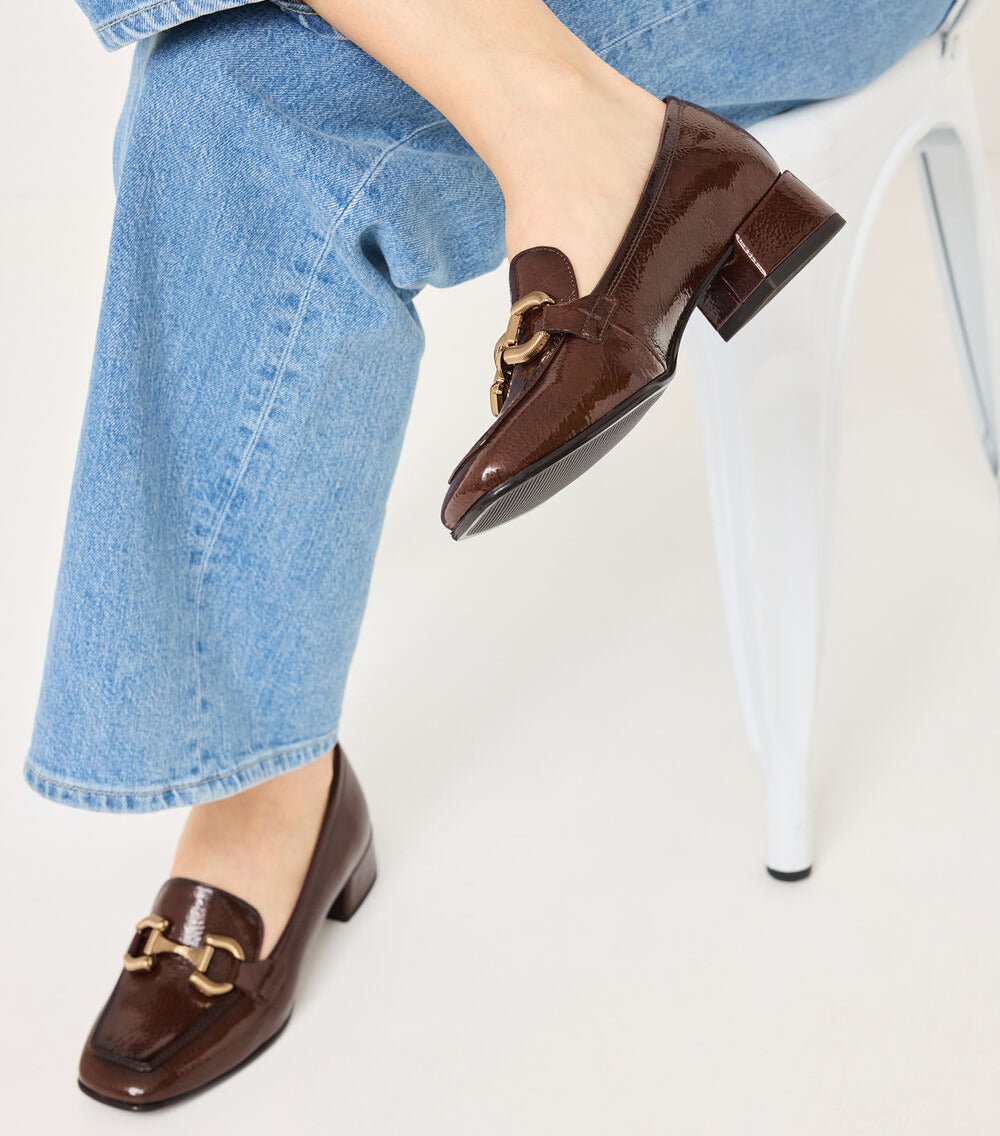 A person wearing brown patent leather loafers with gold-tone horsebit details, paired with light blue jeans, seated on a white chair against a light gray background.