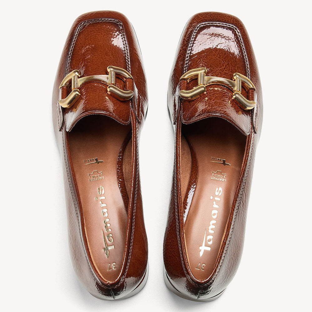 A pair of glossy brown leather loafers with a gold horsebit detail on the toe, shown from a top-down view on a light gray background. The brand name 'Tamaris' is visible on the insole.