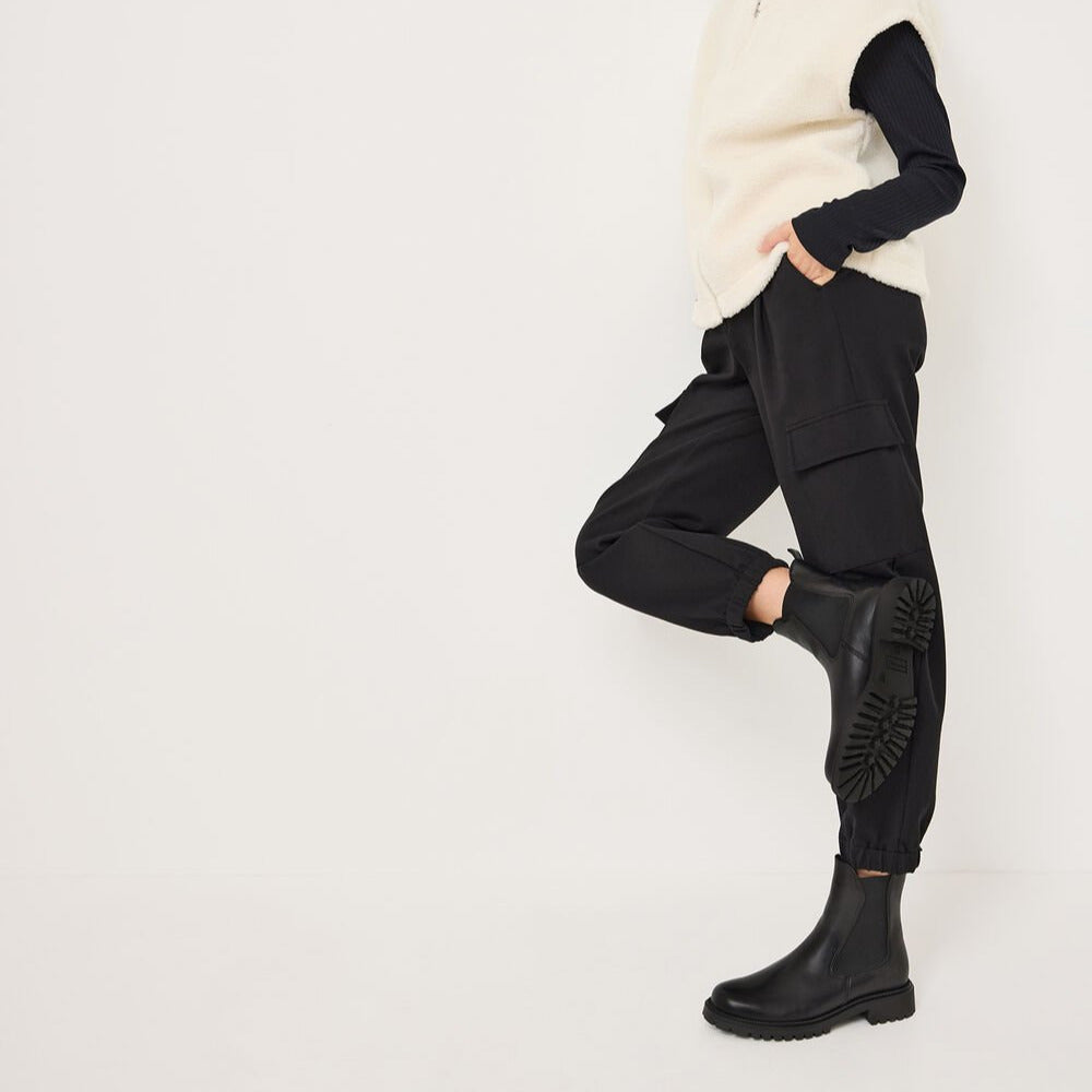 Woman wearing a cream vest, black turtleneck, black pants, and black boots on a white background