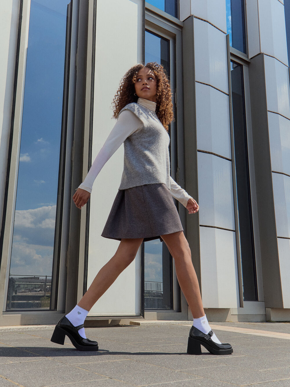 A woman walking outdoors in front of a modern glass building, wearing a light gray knit sweater vest over a white long-sleeve top, a dark gray pleated mini skirt, white socks, and black Mary Jane platform heels.