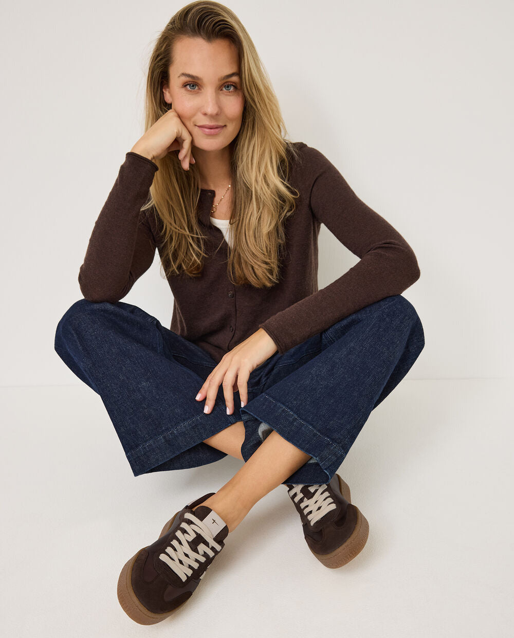 Woman wearing a brown sweater and blue jeans sitting on a white background