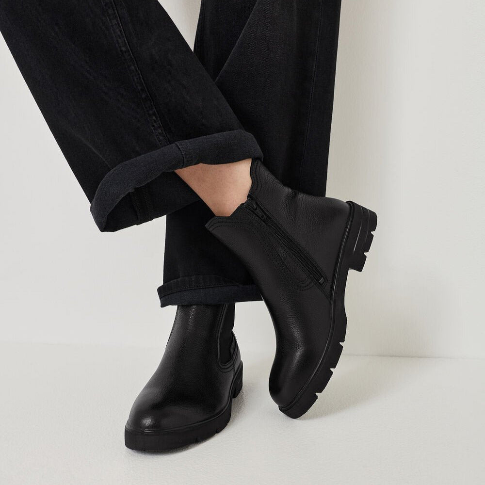 Person wearing black leather boots with rolled-up black pants on a light background