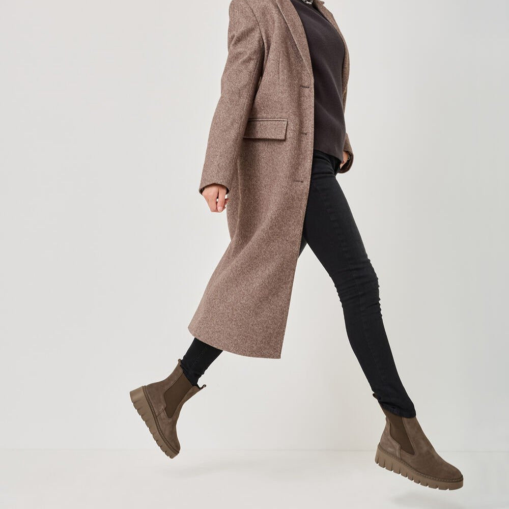 Woman wearing a long brown coat, black turtleneck, black pants, and brown boots on a white background
