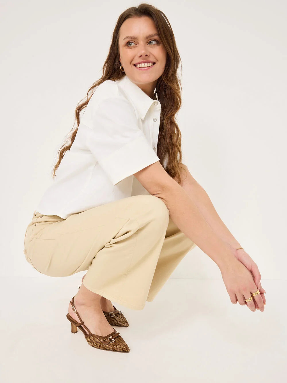 Woman sitting on a white background wearing a white shirt and beige pants.