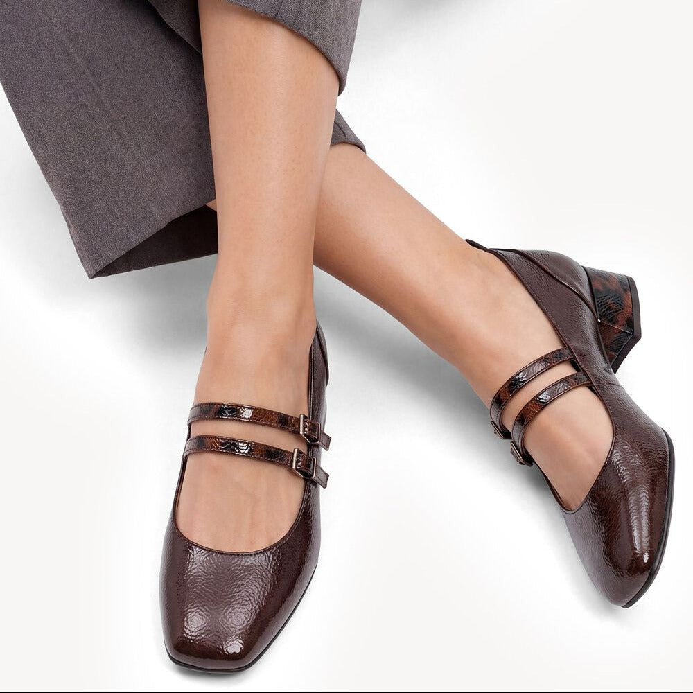 A person wearing dark brown Mary Jane block-heel shoes with double straps and a low block heel, paired with gray trousers, shown against a clean white background.