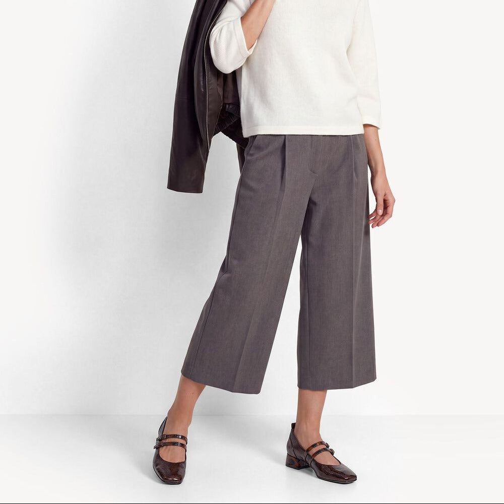 A woman models a white crewneck sweater, gray wide-leg cropped trousers, and dark brown Mary Jane flats, holding a dark brown leather jacket over her shoulder against a clean white studio background.