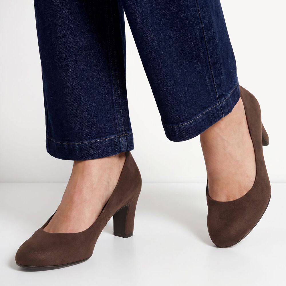 A person wearing dark brown suede-like block-heel pumps with dark blue jeans, shown from the calves down against a clean white studio background.