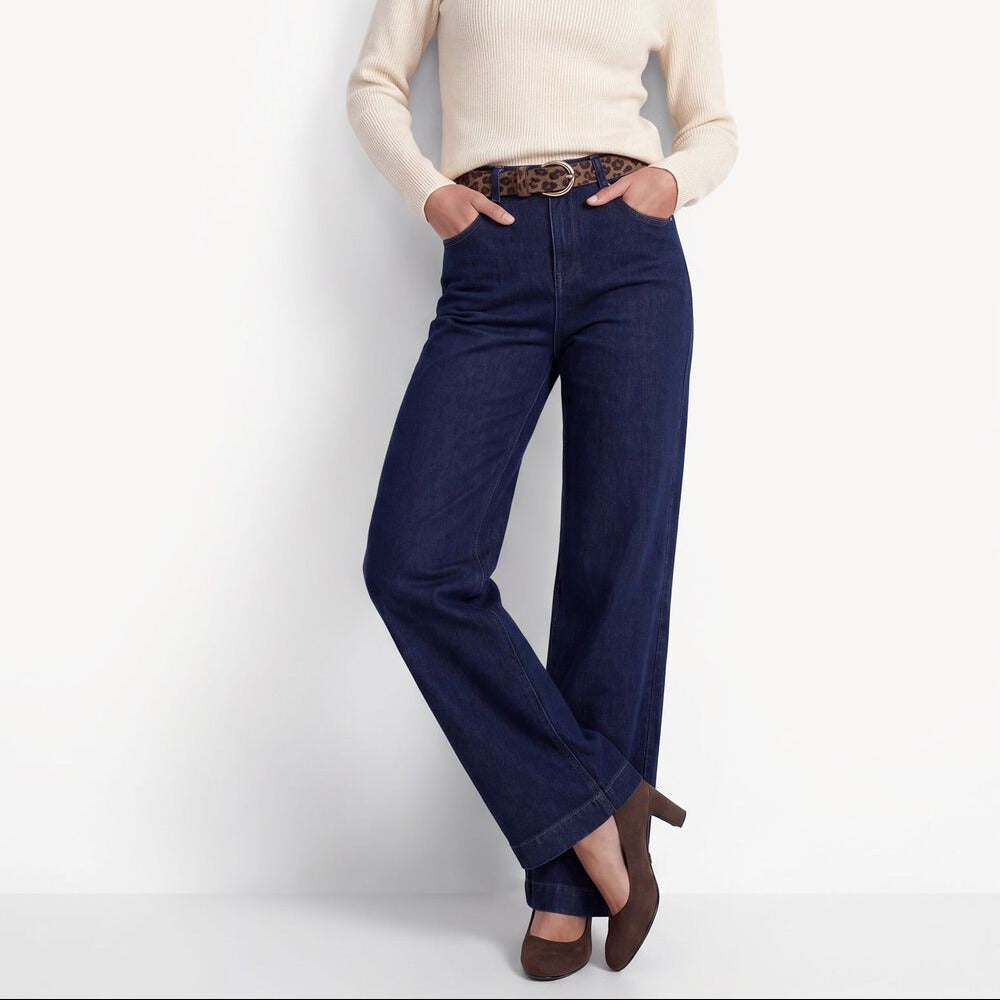A woman modeling a cream mock-neck knit sweater and dark blue wide-leg jeans with a patterned belt, standing against a plain white studio background.