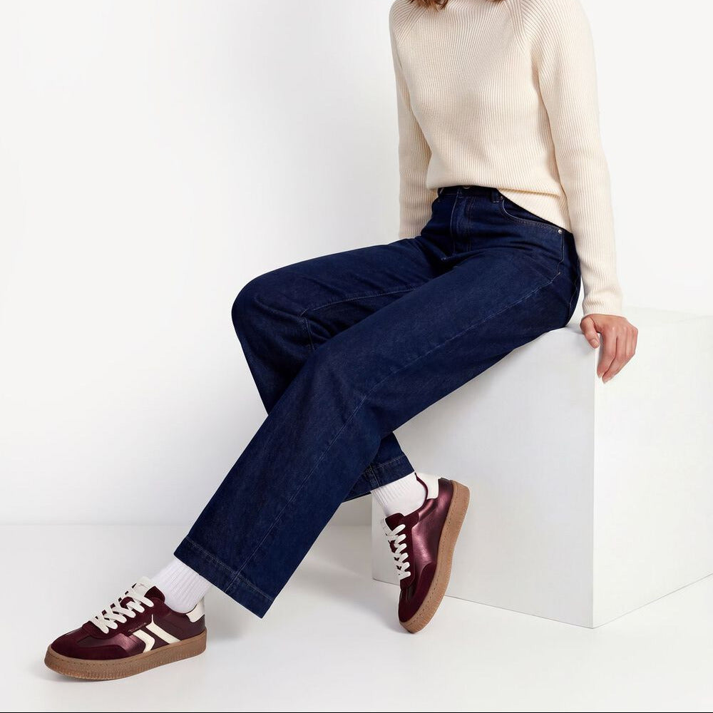 A woman sits on a white block in a studio setting, wearing a cream mock-neck knit sweater, dark blue wide-leg jeans, burgundy sneakers, white socks, and a burgundy beanie.