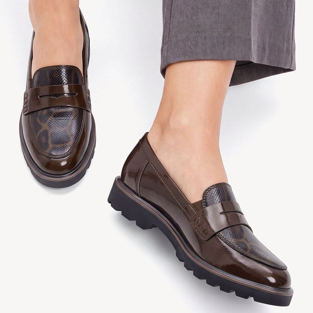 A pair of women's brown leather slip-on loafers with textured snakeskin and smooth leather panels, worn with gray trousers against a white background.