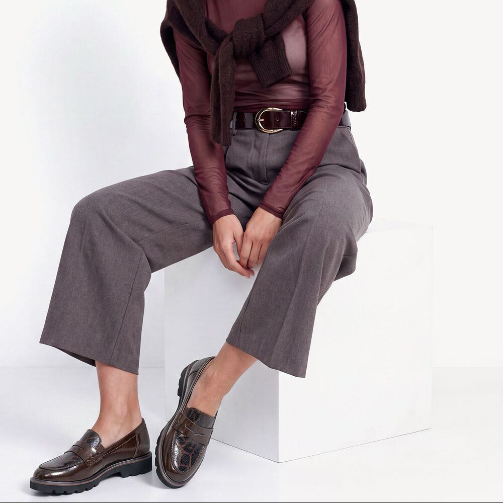 A model sits on a white block wearing a dark brown faux-fur shrug, a sheer burgundy long-sleeve top, gray cropped trousers, and dark brown leather loafers against a white studio background.