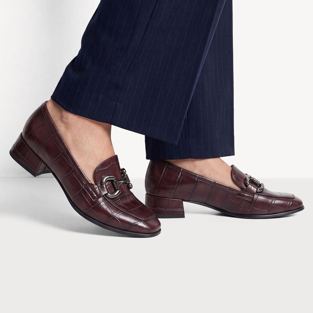 A pair of dark burgundy crocodile-embossed leather loafers with silver-tone horsebit details, shown on a model wearing navy pinstripe trousers against a light gray studio background.