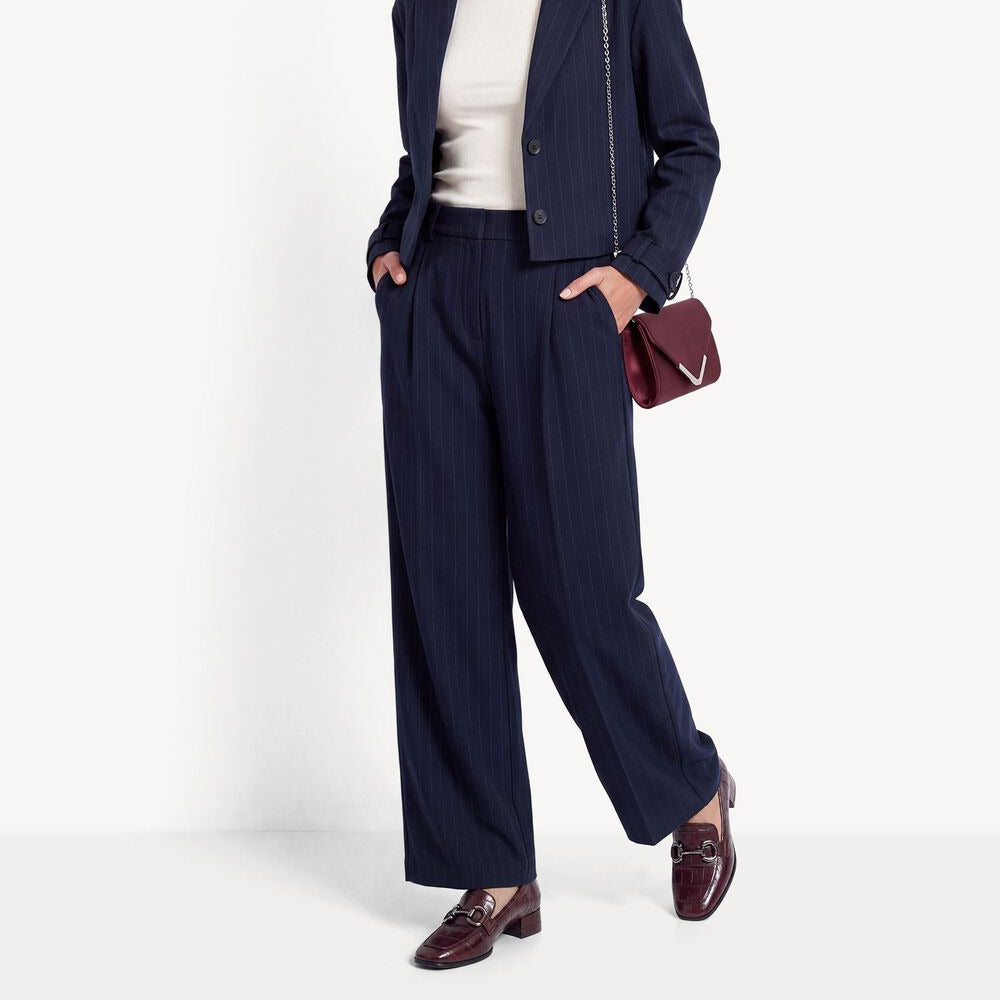 A woman models a navy blue pinstripe blazer and matching wide-leg trousers, styled with a white top, a burgundy handbag, and dark brown loafers against a clean white studio background.