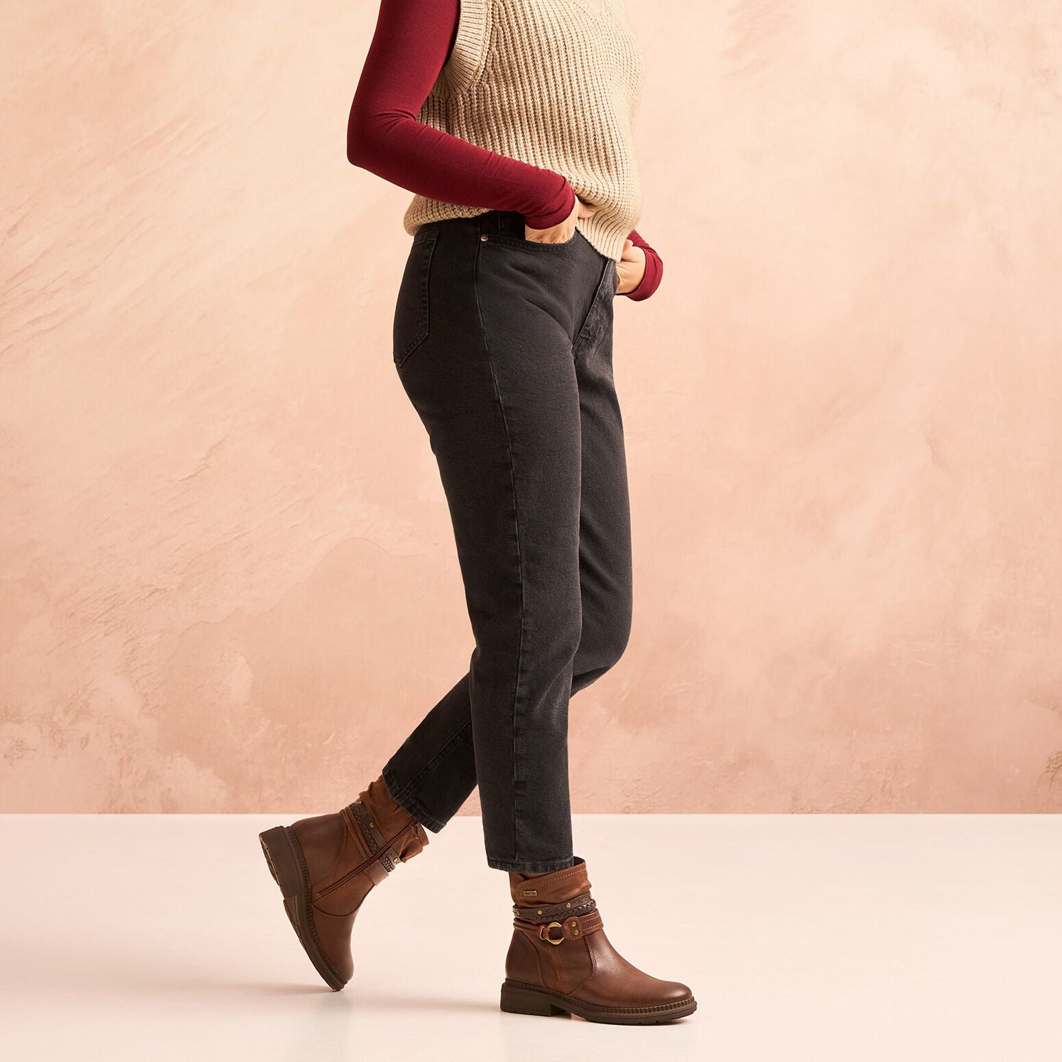 Woman wearing a beige vest, maroon long-sleeve shirt, dark jeans, and brown boots against a pink background
