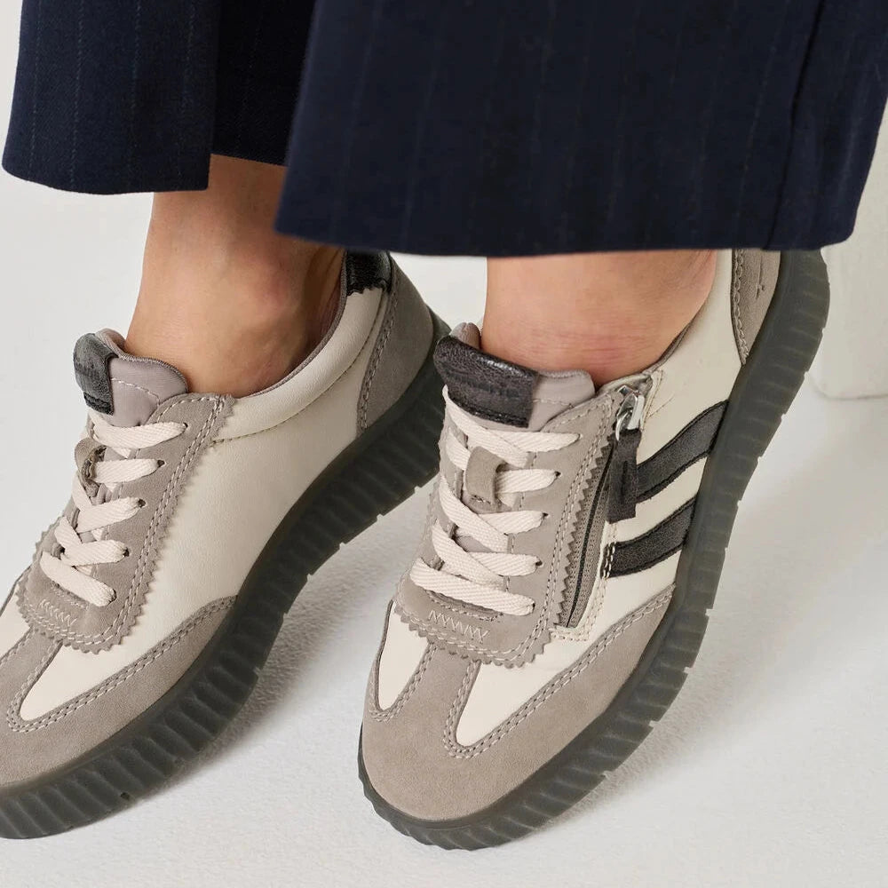 A close-up of a person wearing low-top platform sneakers in taupe, cream, and black, paired with dark navy pinstriped trousers.