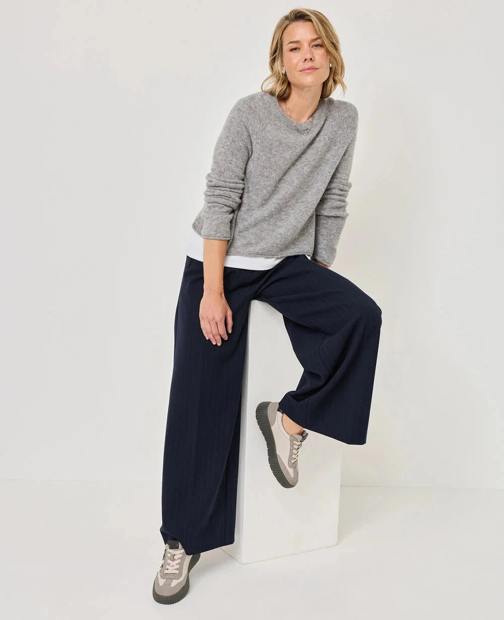 A woman models a gray knit long-sleeve top and navy wide-leg trousers, sitting on a white block against a plain white studio background.