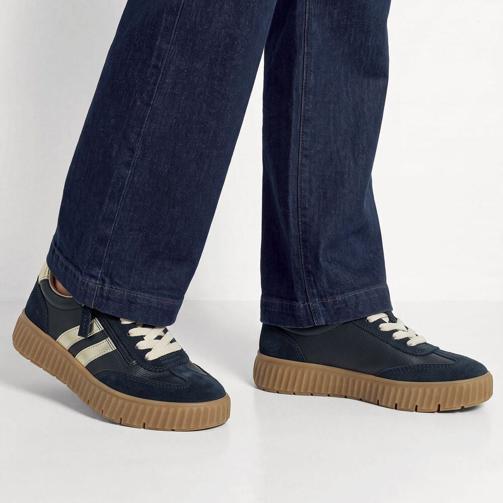 A person wearing dark blue jeans and navy low-top sneakers with cream stripes, white laces, and gum soles, shown from the knees down against a light gray studio background.