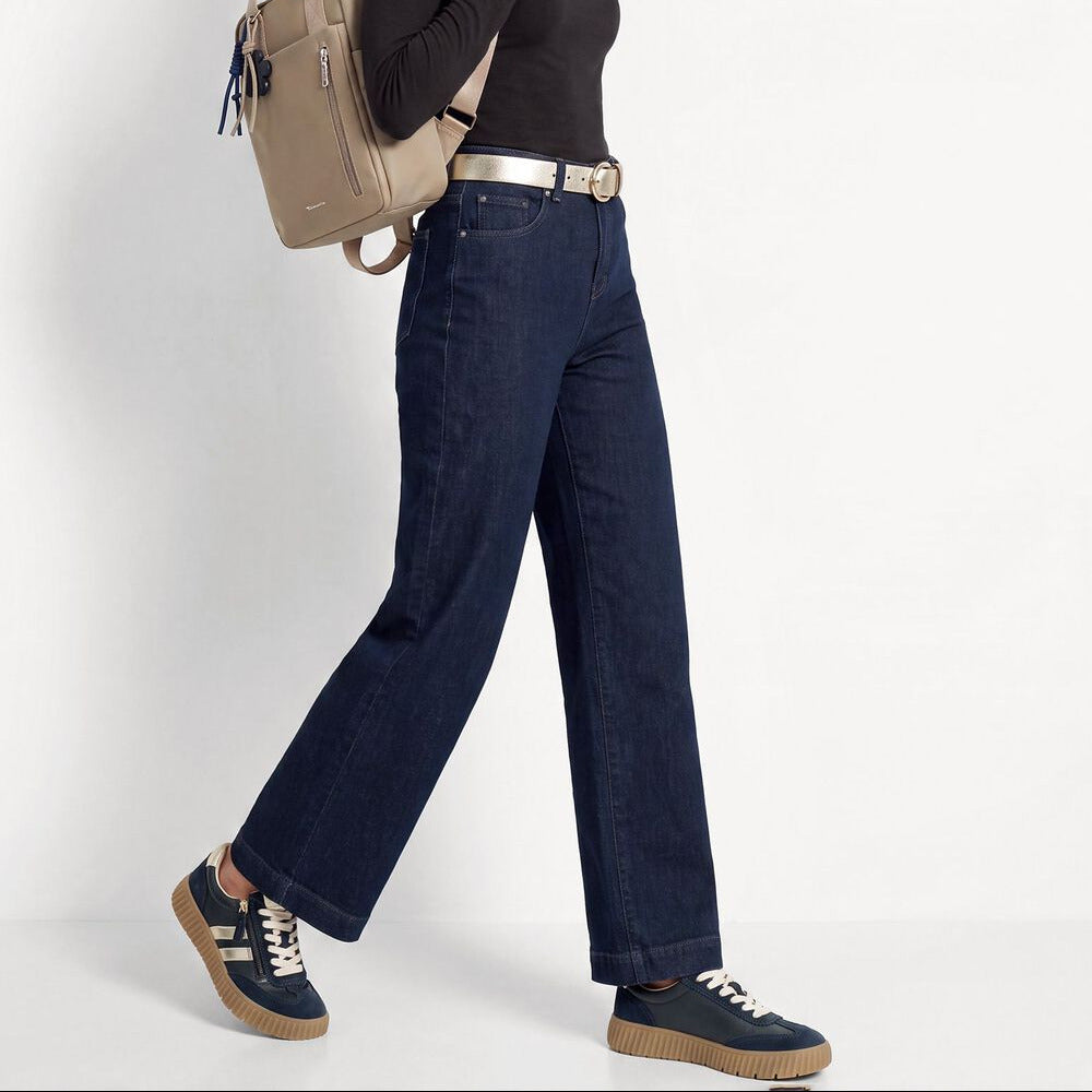 A woman modeling a pair of dark blue wide-leg jeans with a silver belt, paired with a black turtleneck, navy sneakers, and a beige backpack, set against a plain white studio background.