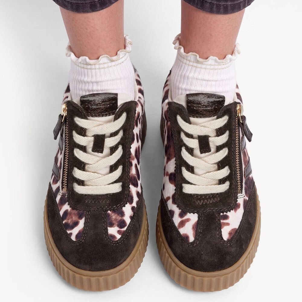 A pair of low-top platform sneakers with a dark brown and pink leopard print pattern, white laces, side zippers, and chunky brown soles, shown on a model wearing white ruffled socks and dark gray jeans.