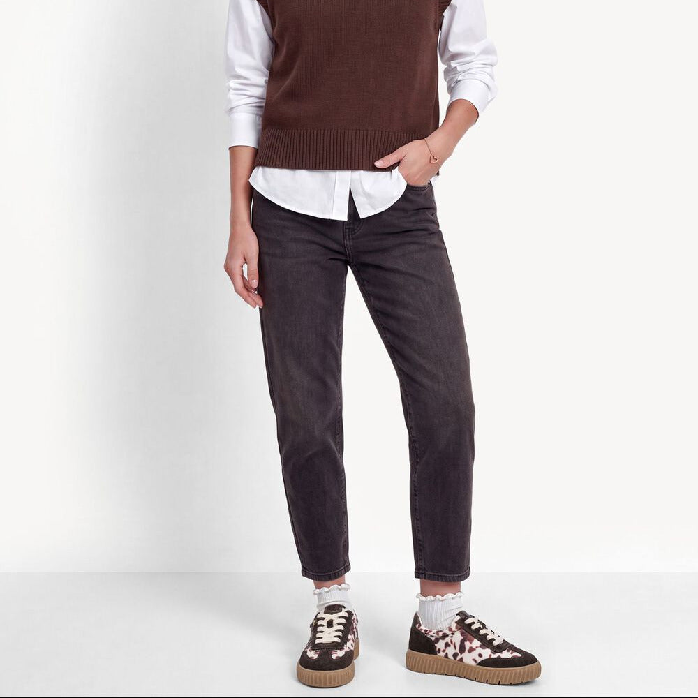 A model stands against a plain white studio background wearing a brown ribbed knit sweater vest over a white collared shirt, dark charcoal gray jeans, and brown-and-white patterned sneakers.