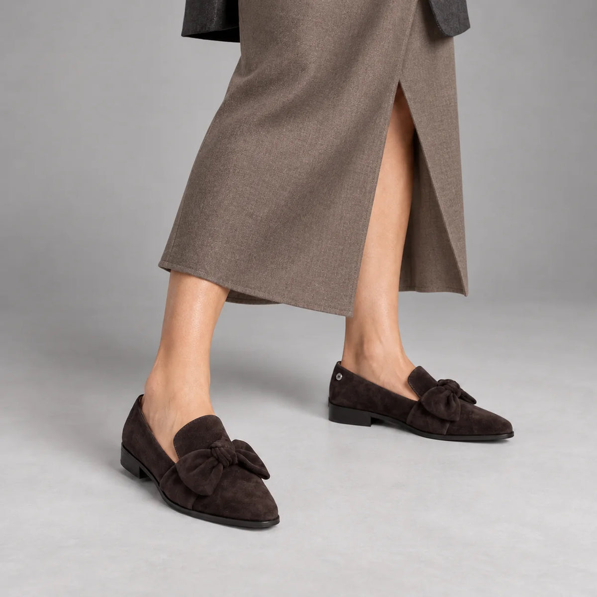 Loretta Vitale W5366 chocolate-brown suede bow loafers worn with a matte taupe tailored midi skirt and bare ankles in a grey studio.