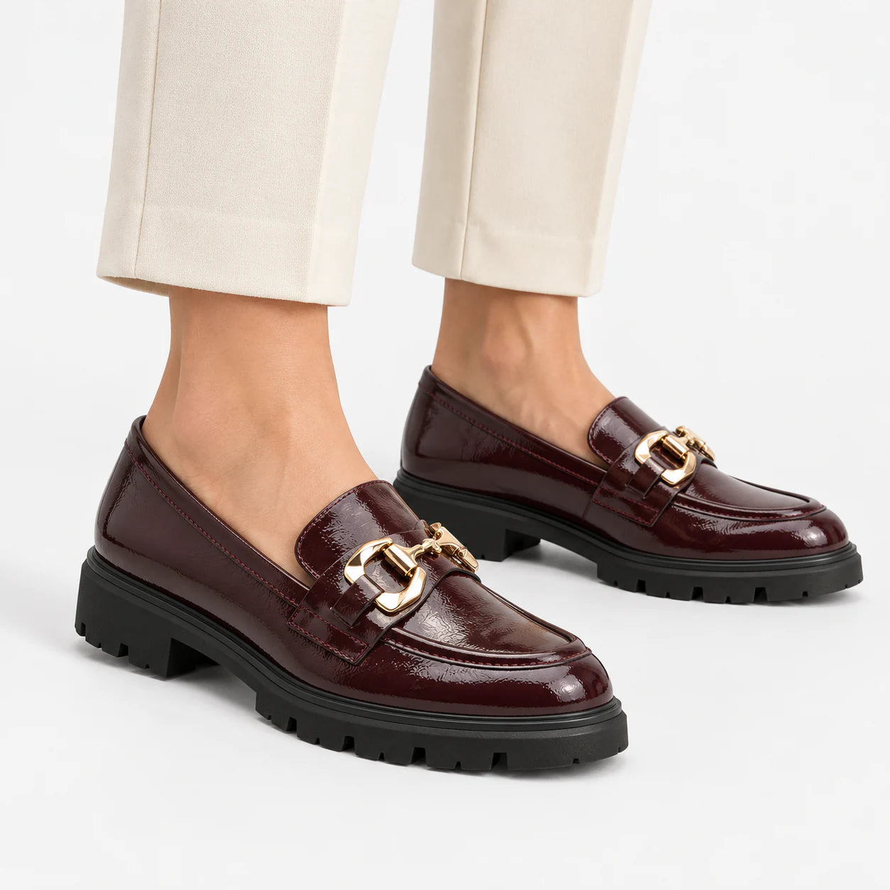 Marco Tozzi Bordeaux patent chunky loafers with gold trim worn with cream trousers.