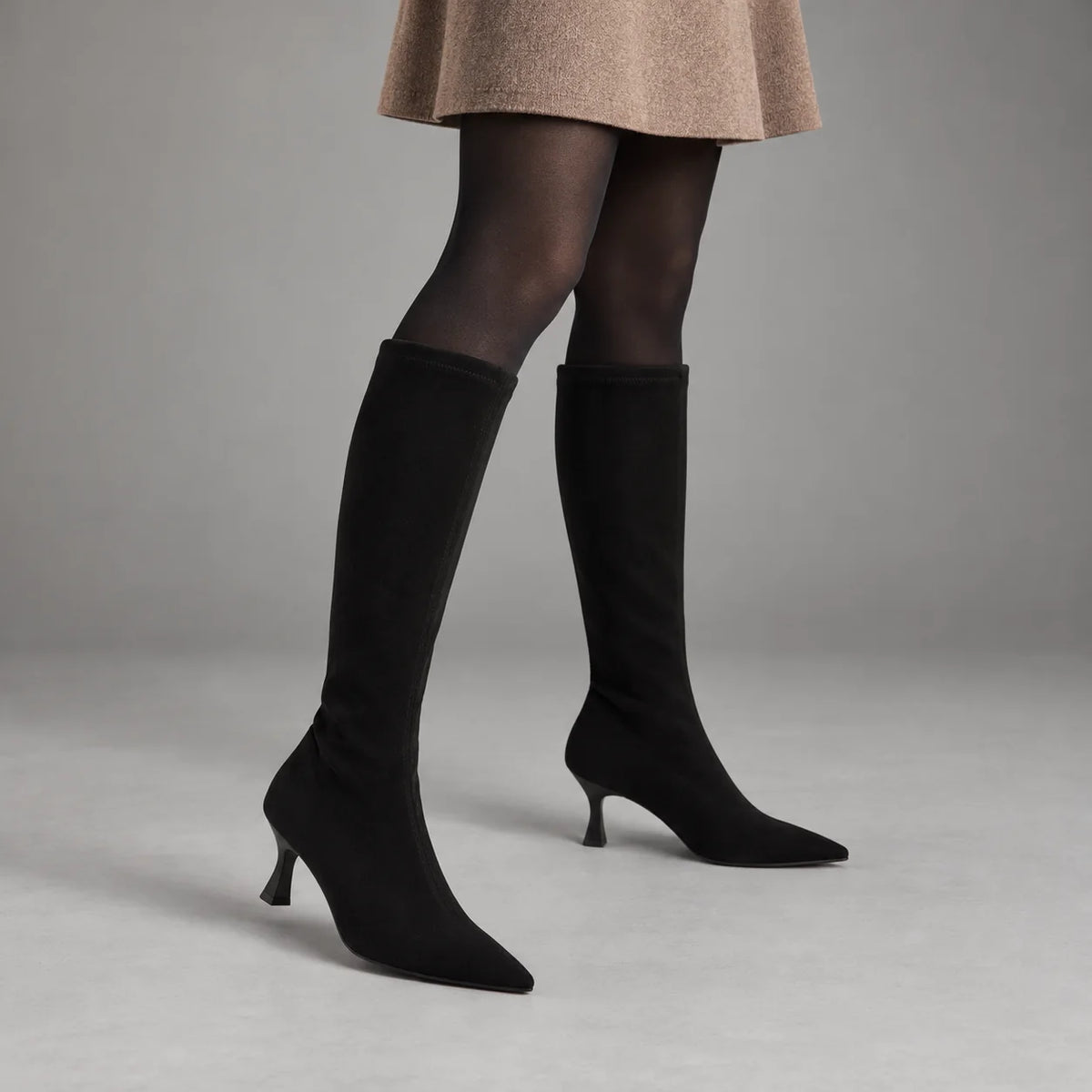 Miss Elastic Zeta black stretch knee-high boots styled with a camel skirt and black tights in a grey studio
