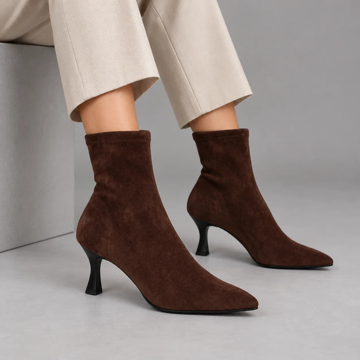 Model wearing Miss Elastic Zeta brown stretch ankle boots with ivory cropped trousers, seated in a grey studio