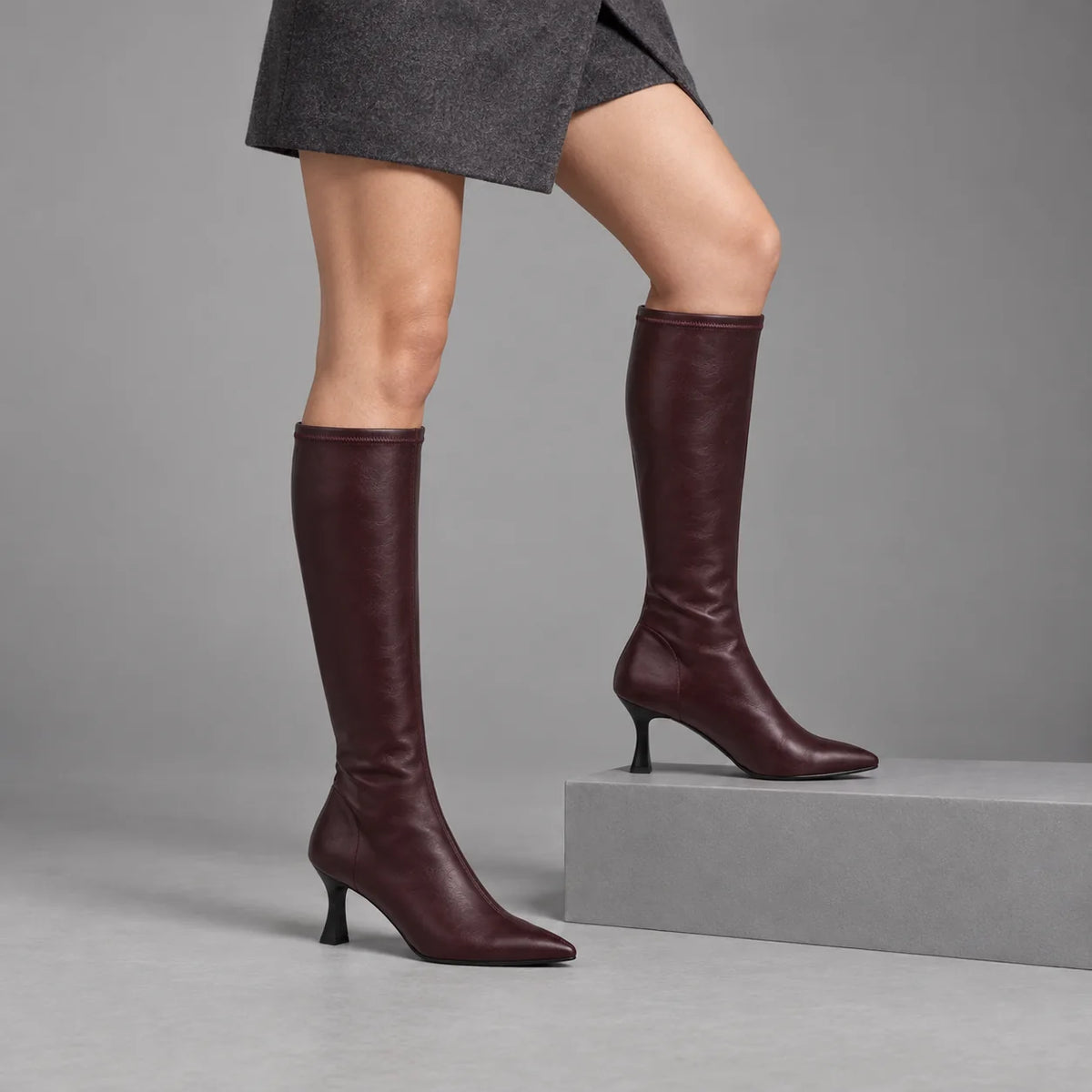 Miss Elastic Zeta burgundy knee high boots worn with a charcoal skirt, one foot on a low plinth in a grey studio.