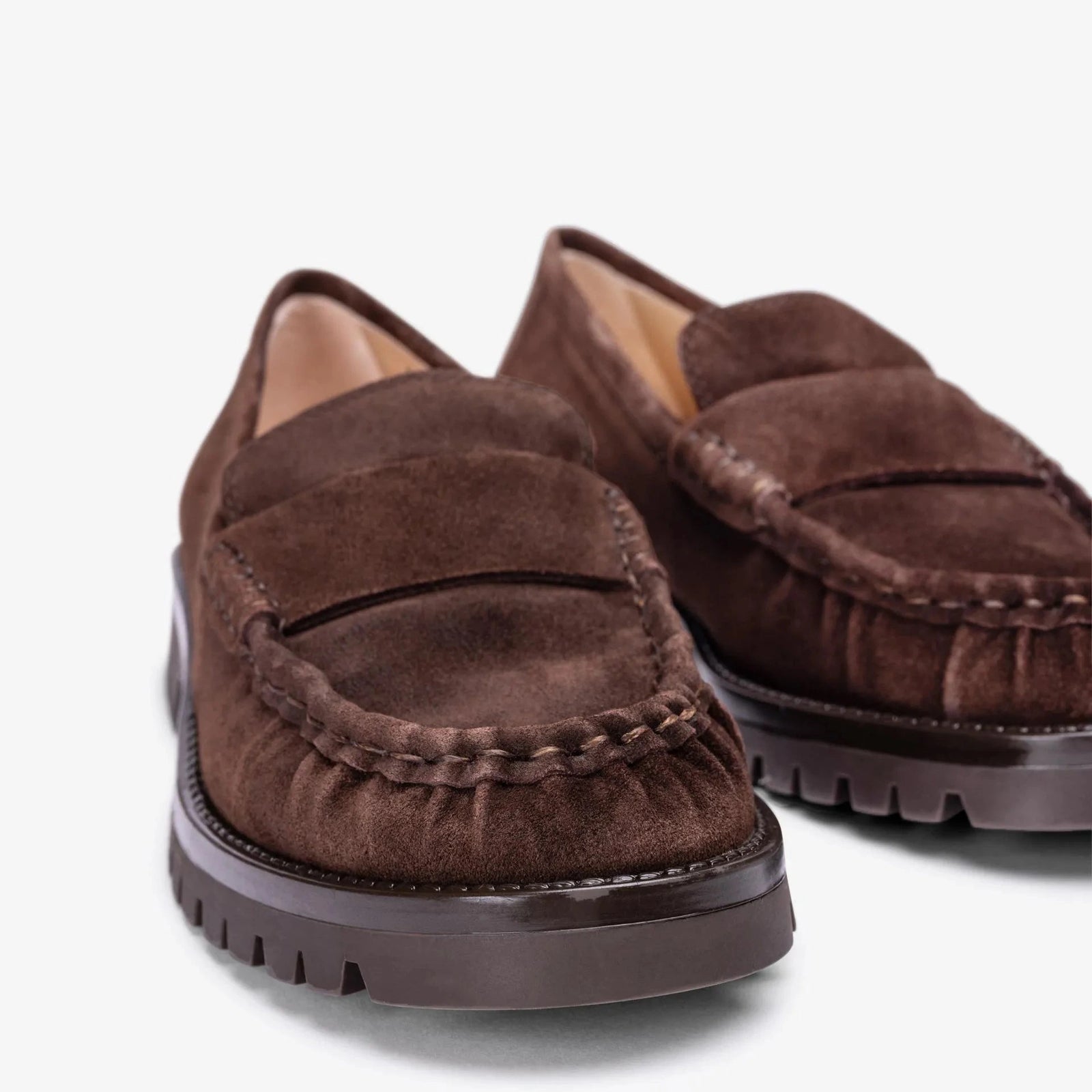 A pair of dark brown suede penny loafers with thick dark brown lug soles, shown from a high-angle perspective on a clean white background.