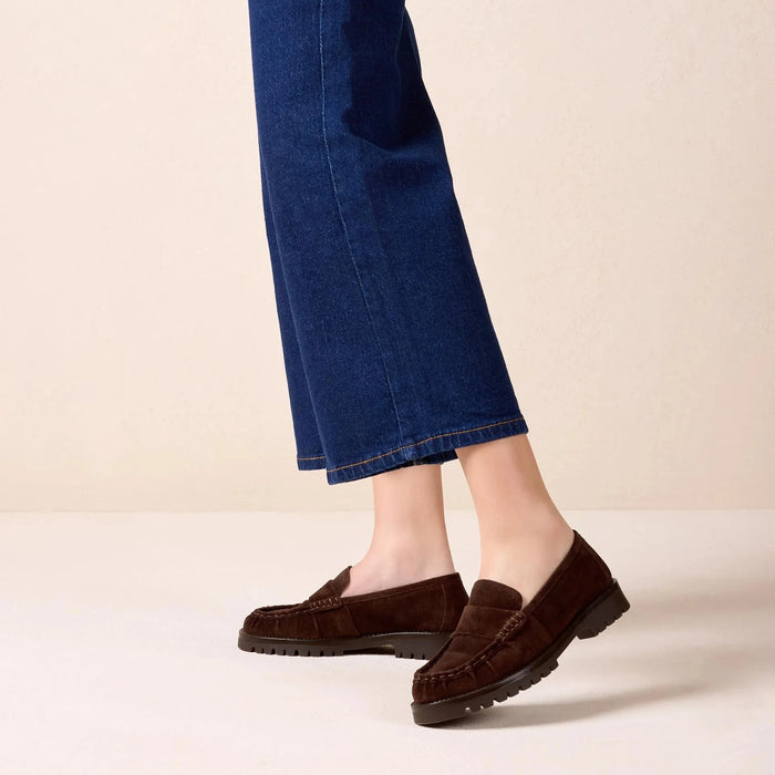 A person wearing dark brown suede moccasin loafers with dark soles, paired with blue cropped jeans and a navy and gold striped blazer, standing against a solid beige studio background.