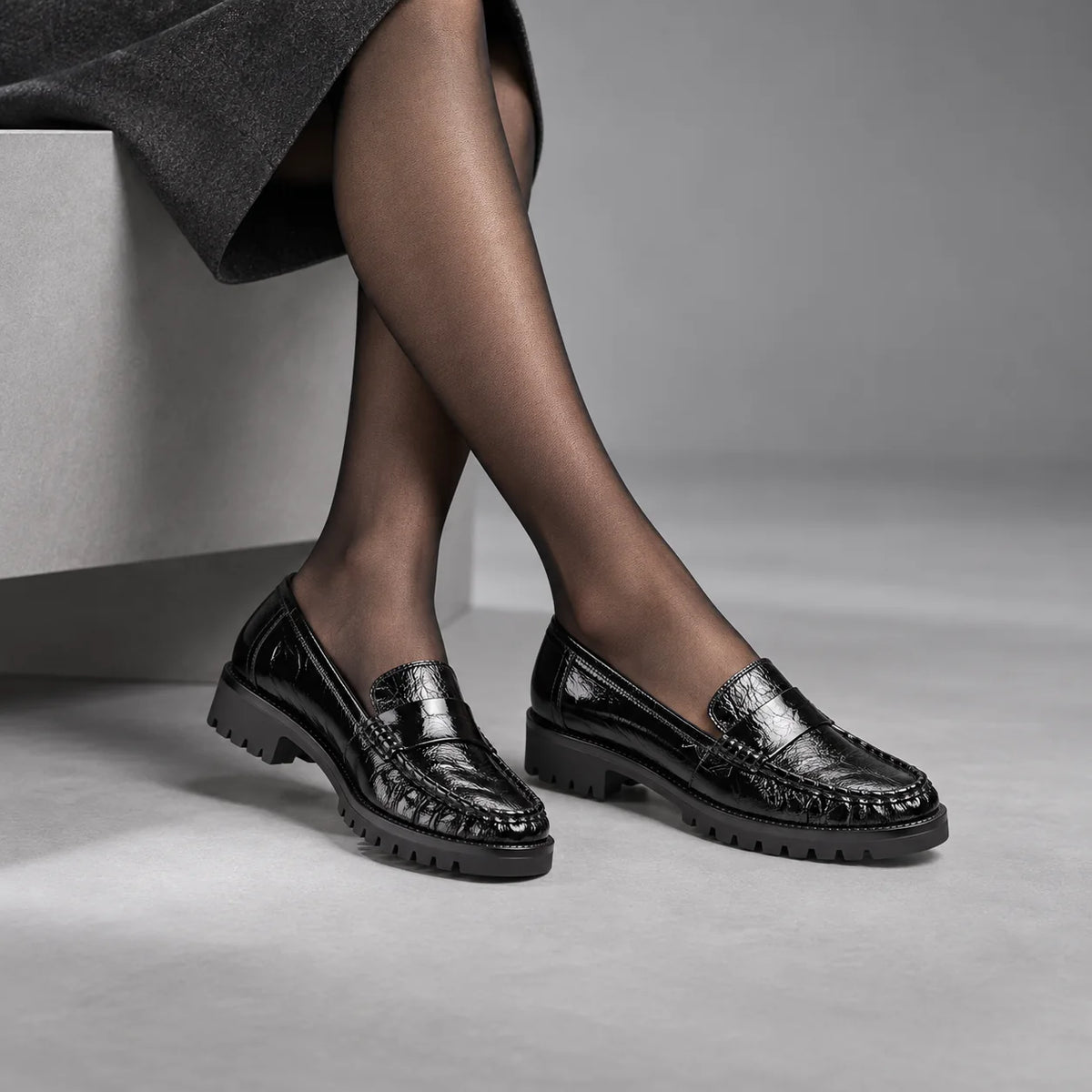 Unisa Fijo black patent leather loafers worn with sheer tights and a charcoal skirt in a grey studio.